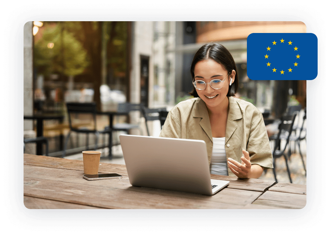 Young woman with glasses working on a laptop at an outdoor café, with the European Union flag in the top right corner.